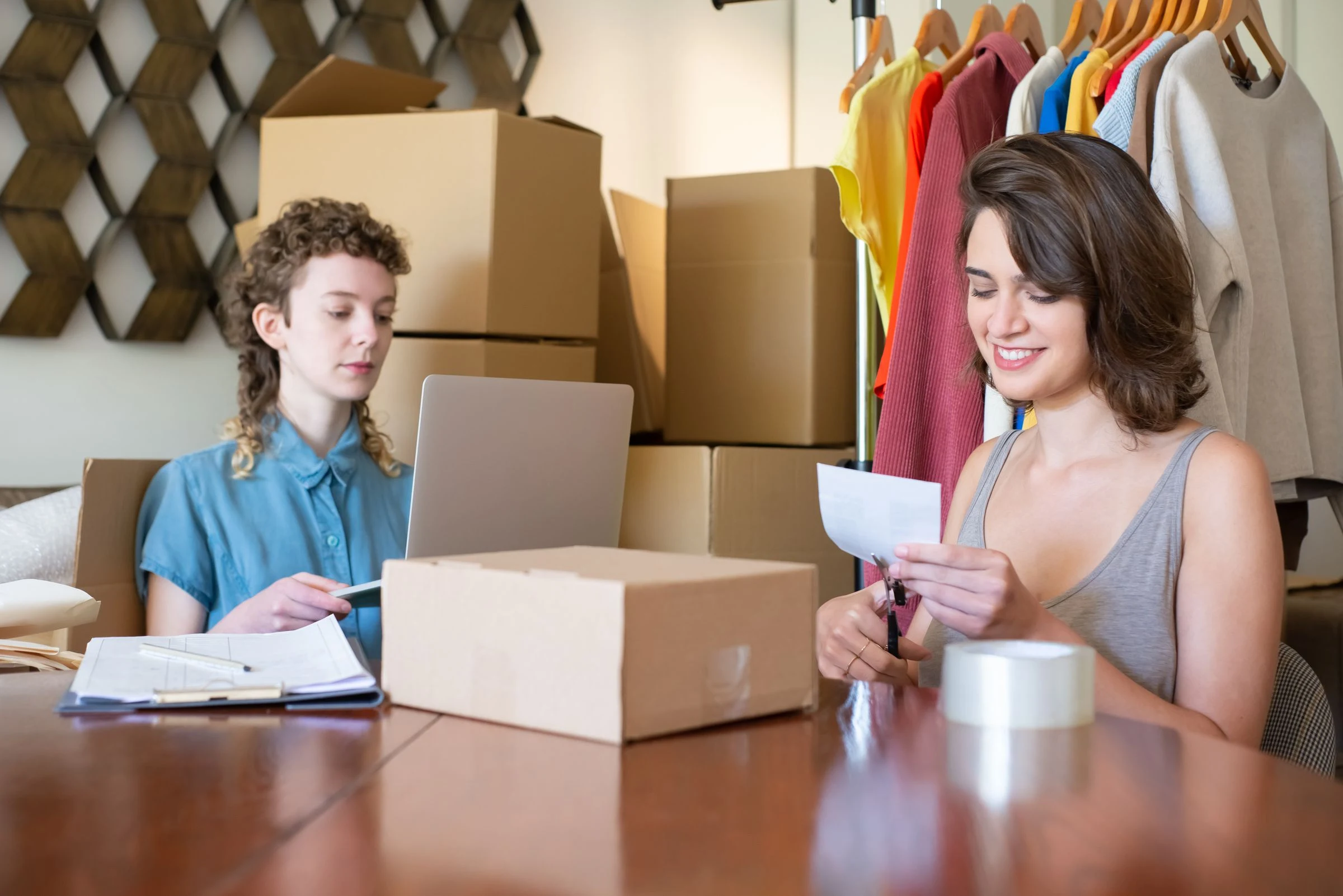 Dos personas preparan pedidos de una tienda online junto a una computadora, prendas y materiales de empaque.