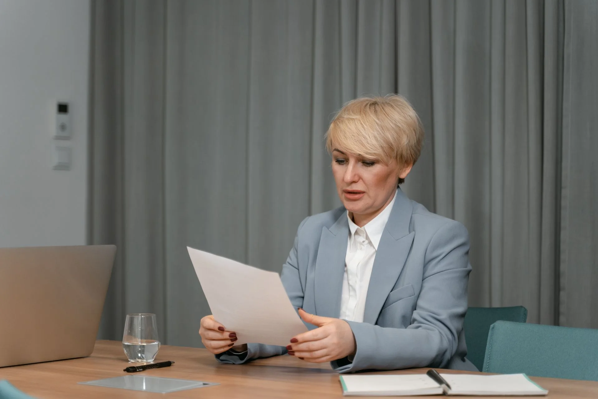 A professional in a blazer reviews printed documents at a desk beside a laptop.